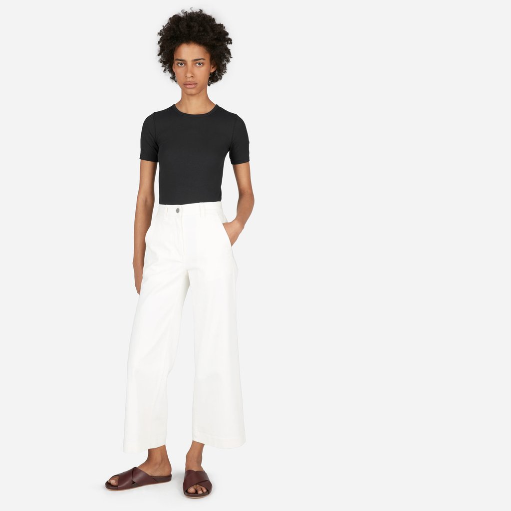 A woman in an black Everlane t-shirt and white pants stands against a white background.