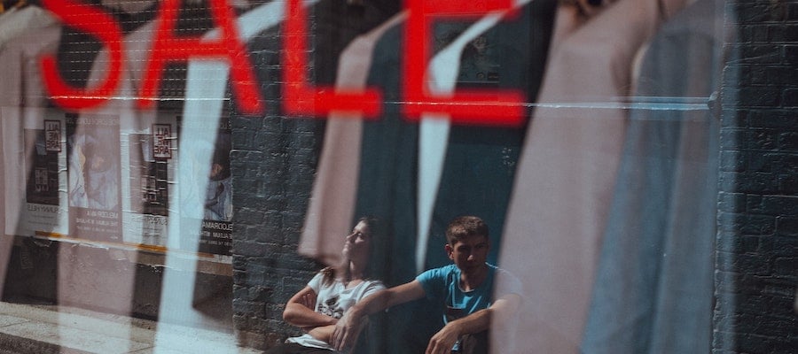Two seated people reflected in the glass of a storefront with a red sale sign, by Clem Onojeghuo.
