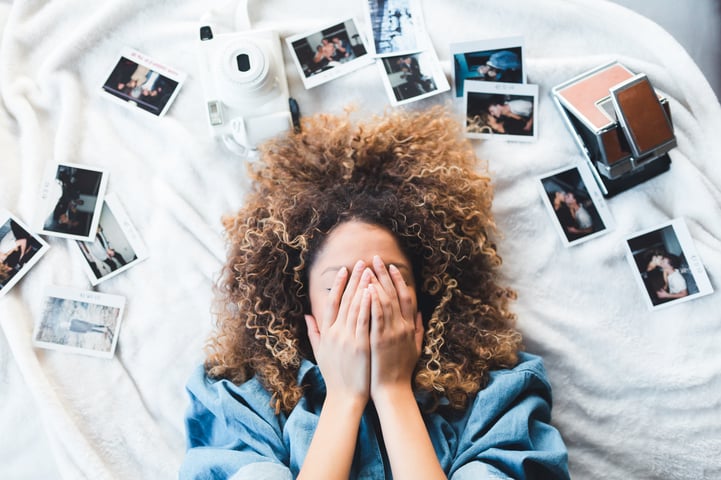 Woman lying on bed covering her face while surrounded by Polaroid pictures.