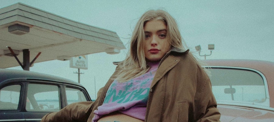 An influencer in a brown jacket seated on the hood of a car, by Joshua Rondeau.