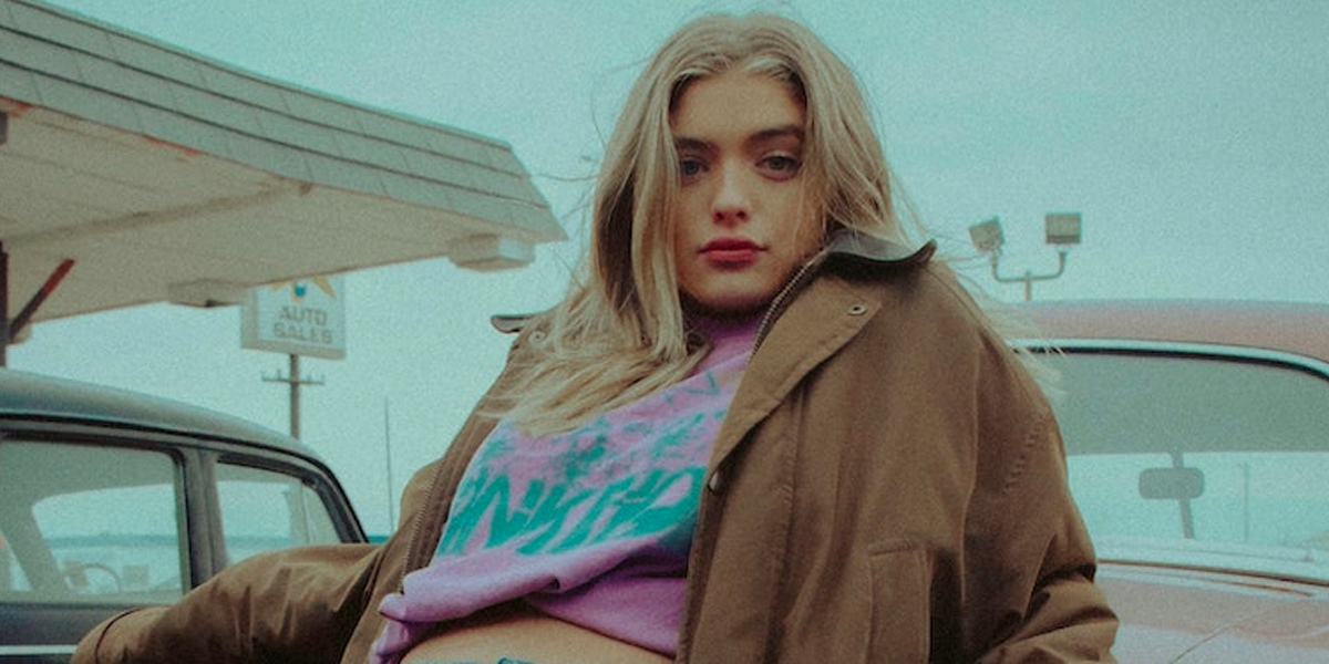 An influencer in a brown jacket seated on the hood of a car, by Joshua Rondeau. 