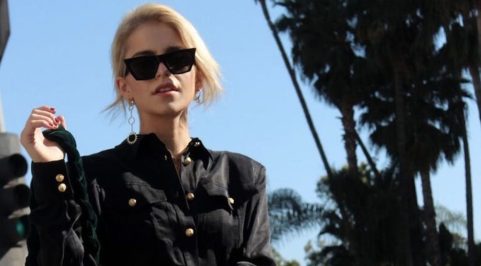 Caroline Daur walks past palm trees in black sunglasses and a leather jacket.