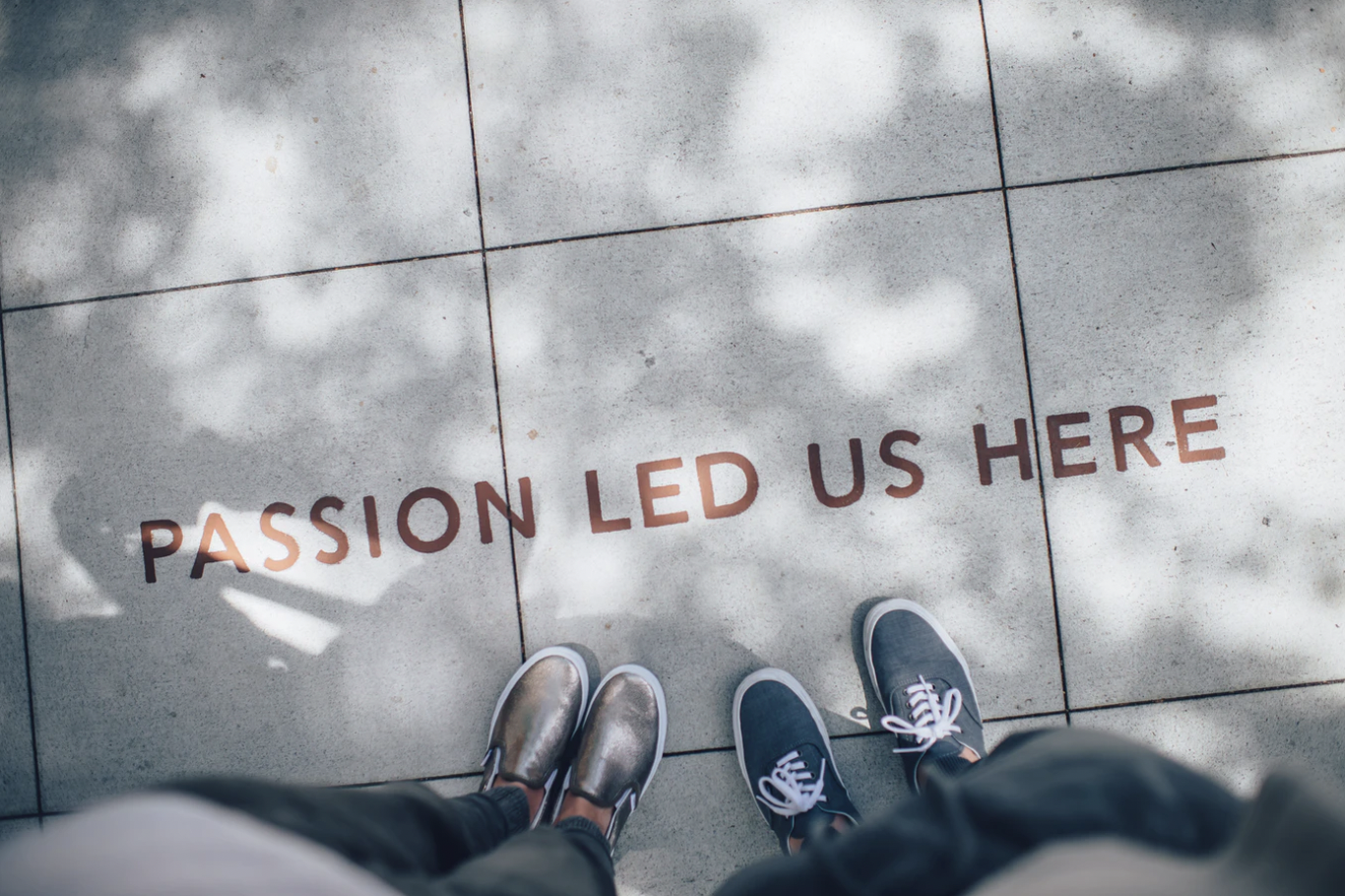 A photo by Ian Schneider, featuring the shoes of two individuals standing next to a sign on the ground that reads “Passion Led Us Here.” 
